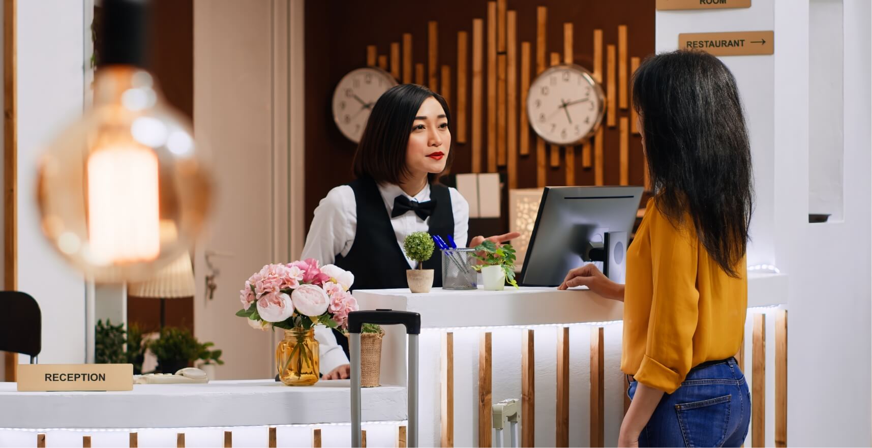 Hotel reception scene: a receptionist in a white shirt, black vest, and bow tie assists a guest at the front desk, flowers on the counter nearby.