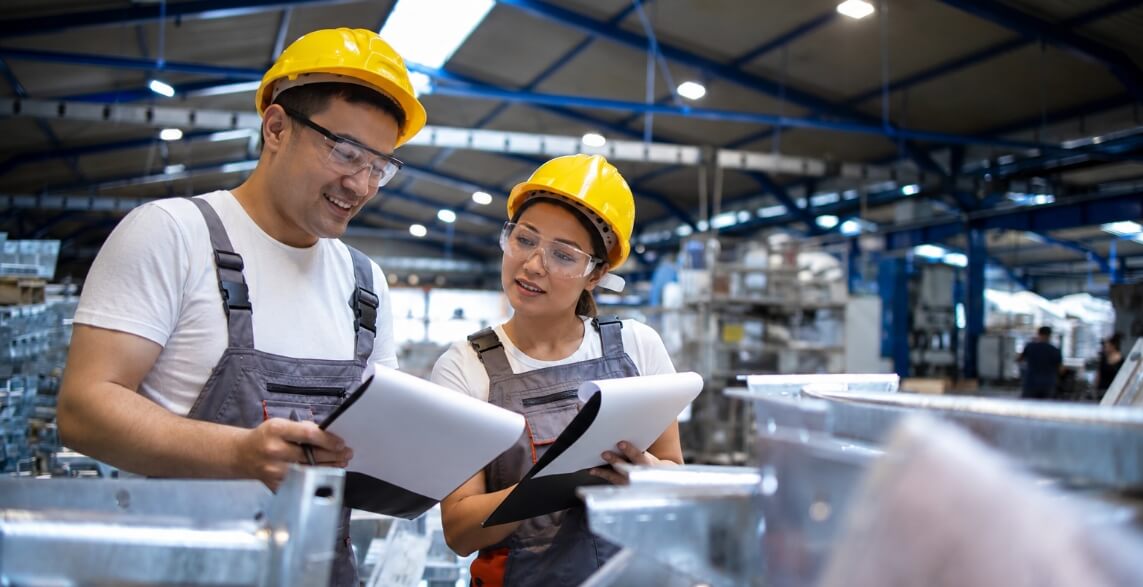 Two factory workers in yellow hard hats and safety goggles review documents together in a busy industrial setting.