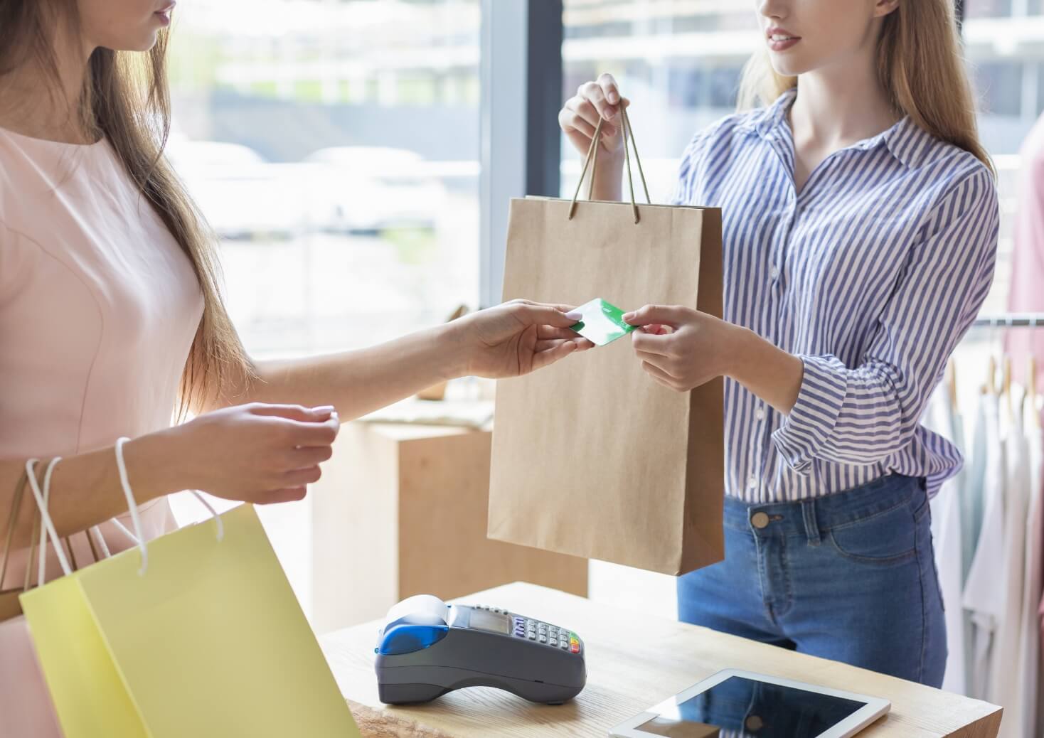 Customer hands a card to a store clerk at the checkout, with a brown paper bag on the counter and a payment terminal nearby.