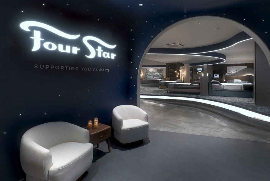 Showroom entrance with a curved lit arch leading to a bed display; navy wall features the Four Star logo and 'Supporting you always' text.