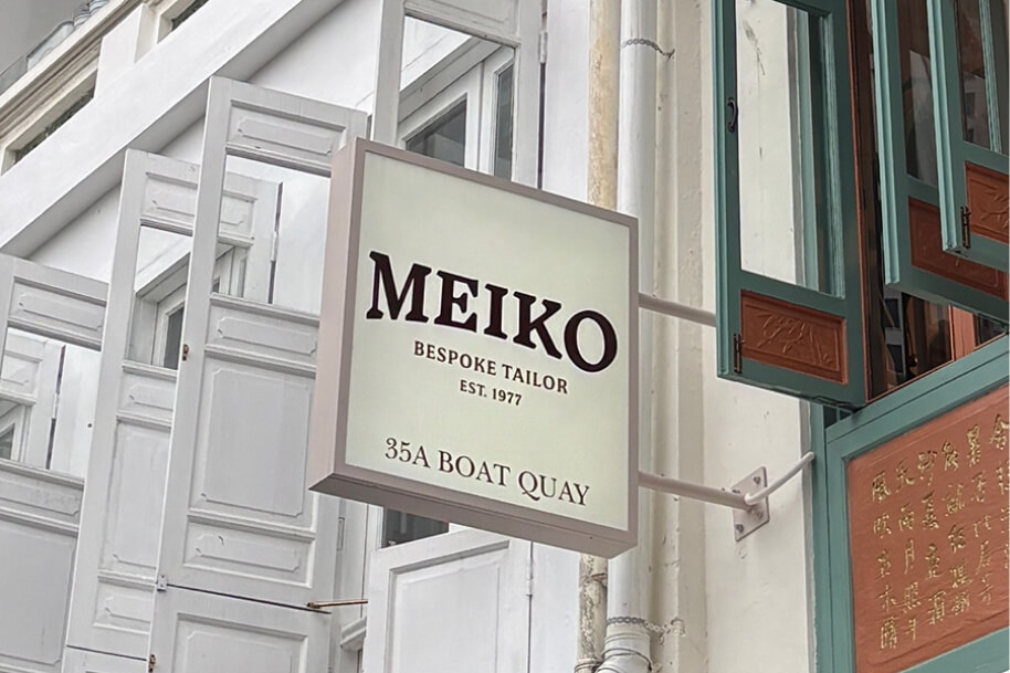 Square hanging storefront sign reads 'MEIKO' with smaller text 'Bespoke Tailor', 'EST. 1977', and '35A Boat Quay' against a white building with teal-green window frames.