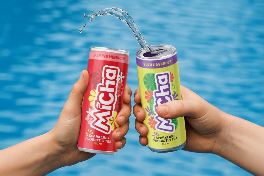 Two hands hold pink and lime-green cans of sparkling probiotic tea labeled Jasmine Hibiscus and Yuzu Lavender, with a splash between them against a blue background.
