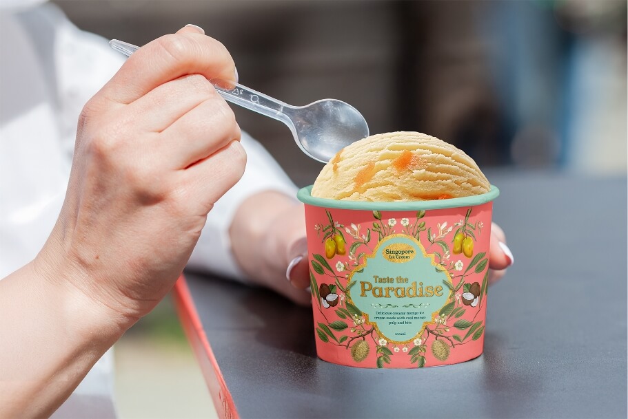 Close-up of a hand with a spoon about to scoop ice cream from a pink Paradise-branded cup with fruit illustrations.