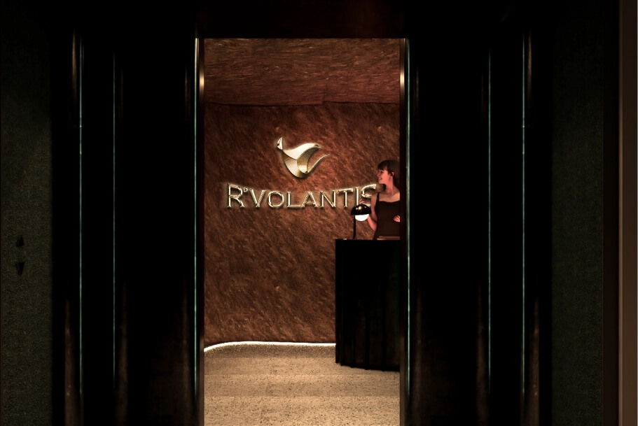 Reception area with a woman at a black podium in front of a bronze textured wall featuring a logo and brand name behind her.