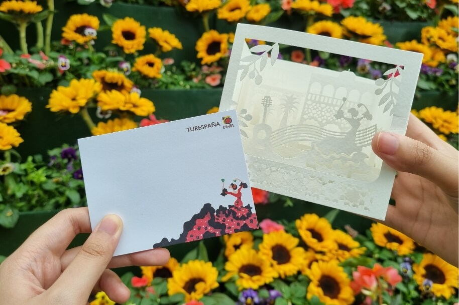 Two hands hold two decorative cards in front of a vibrant flower bed with yellow sunflowers behind they frame.
