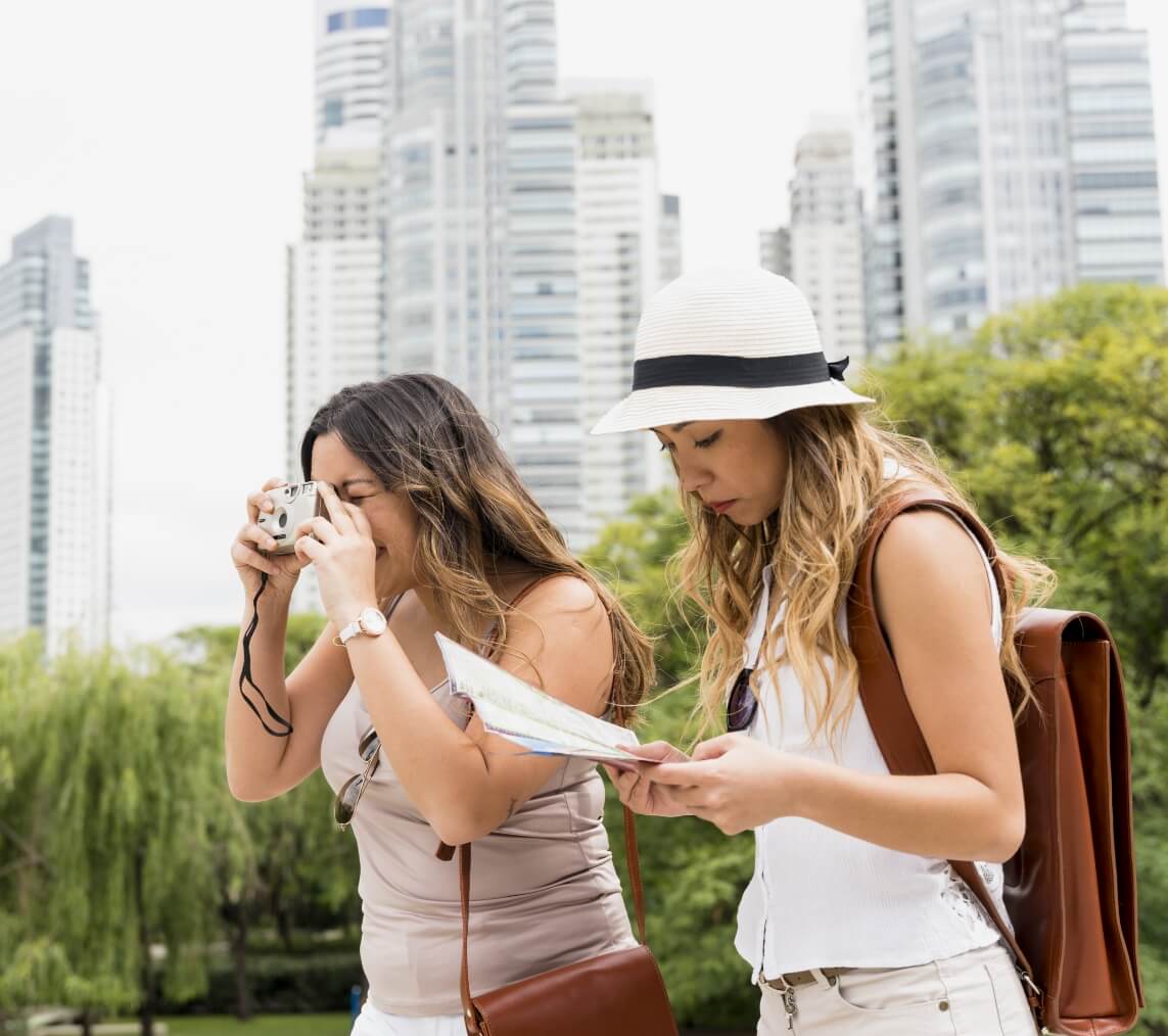 Two women tourists in a city park; one uses a camera while the other reads a map or brochure together.