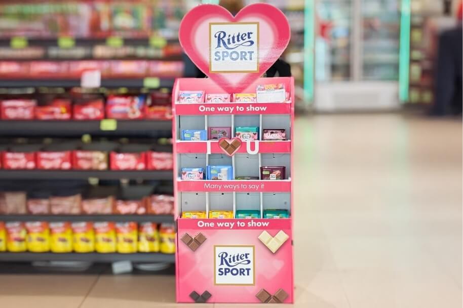 Promotional Ritter Sport chocolate display in a pink stand with a heart top in a supermarket, showcasing assorted chocolate bars.