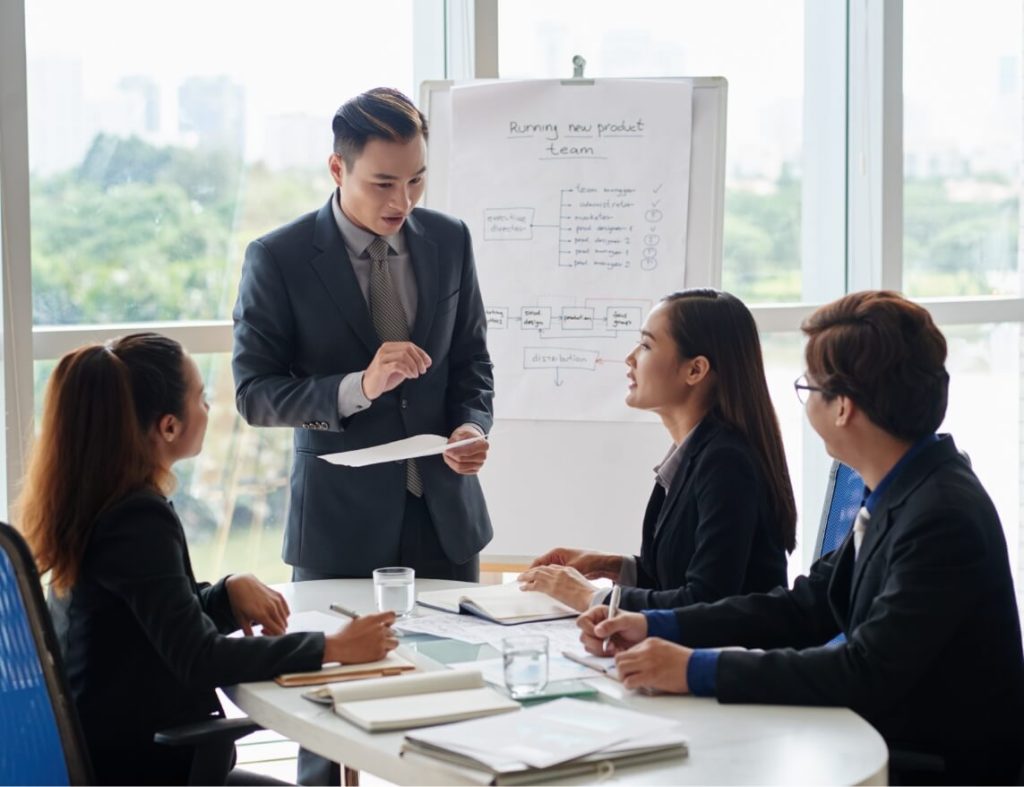 A business meeting with four people around a table as a man in a suit presents beside a flip chart titled 'Running new product team'.
