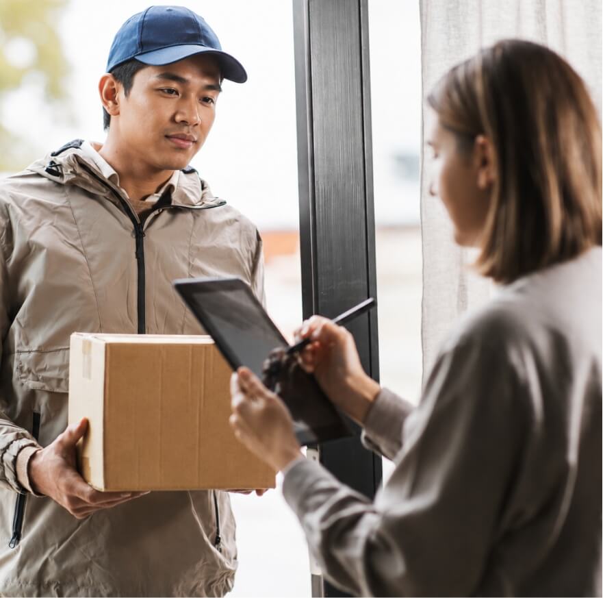 Delivery person in a blue cap hands a cardboard package to a customer at a doorway, who signs on a tablet.