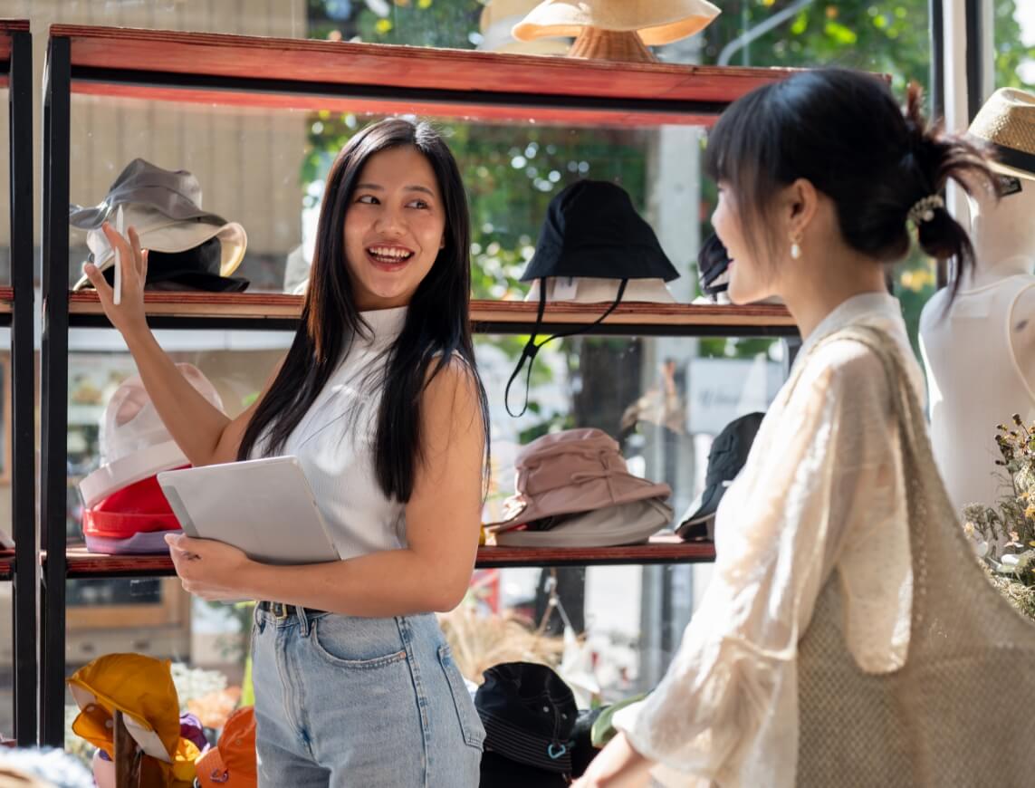 Woman with long black hair holds a tablet and smiles while pointing at hats on a display shelf with another shopper nearby.