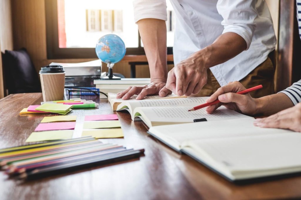 Two people study at a wooden desk with an open book, a red pencil, colorful sticky notes, and a takeaway coffee cup nearby.