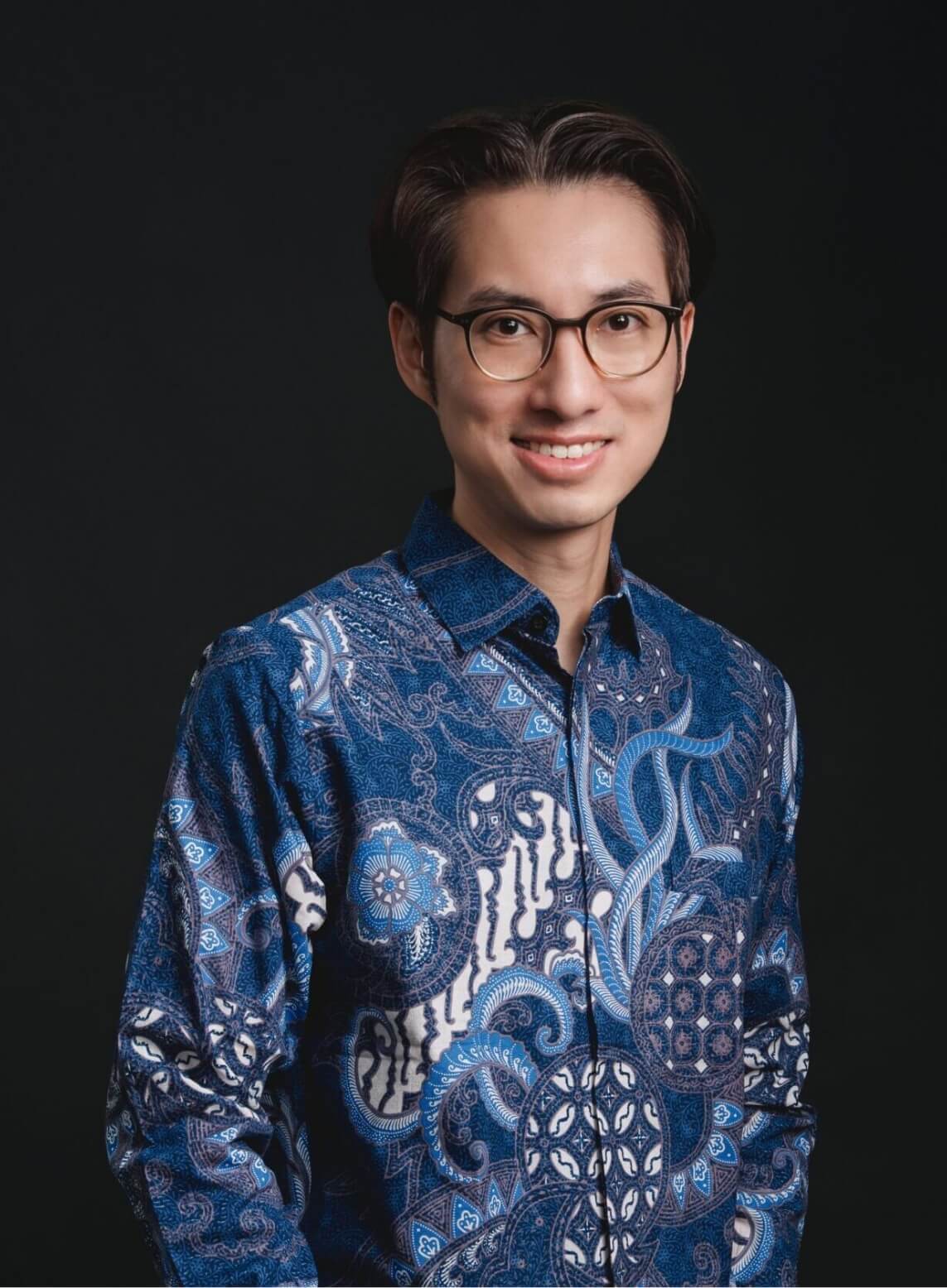 Portrait of a smiling man with glasses wearing a blue batik shirt against a dark backdrop.