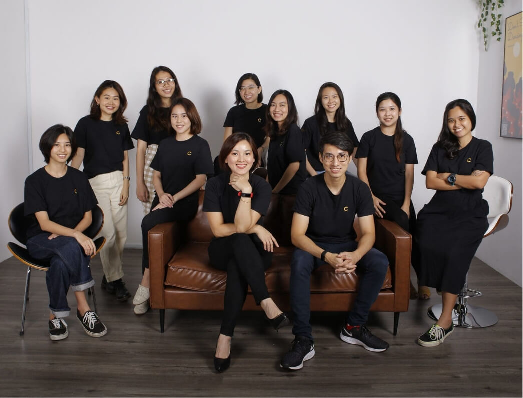 Twelve coworkers in black shirts with a small yellow logo pose in a modern office around a brown leather sofa.