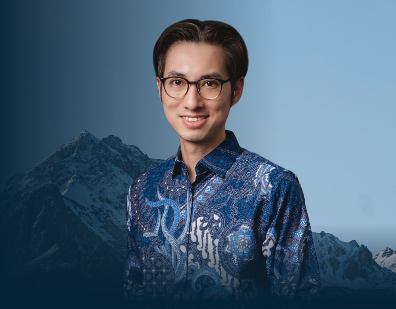 Portrait of a smiling man with glasses in a patterned blue shirt, with mountains in a blue-toned background behind him.