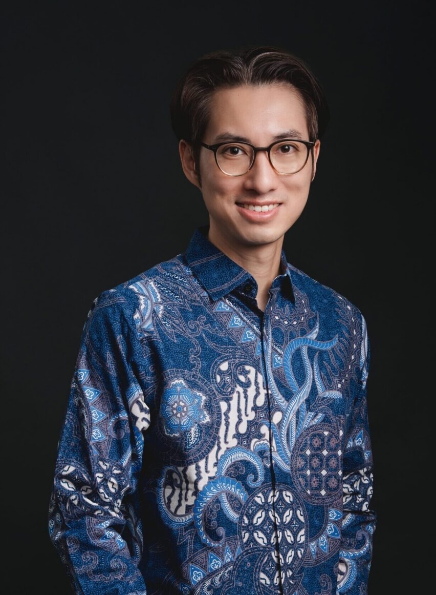 Portrait of a man with glasses, smiling, in a blue patterned shirt against a dark background.