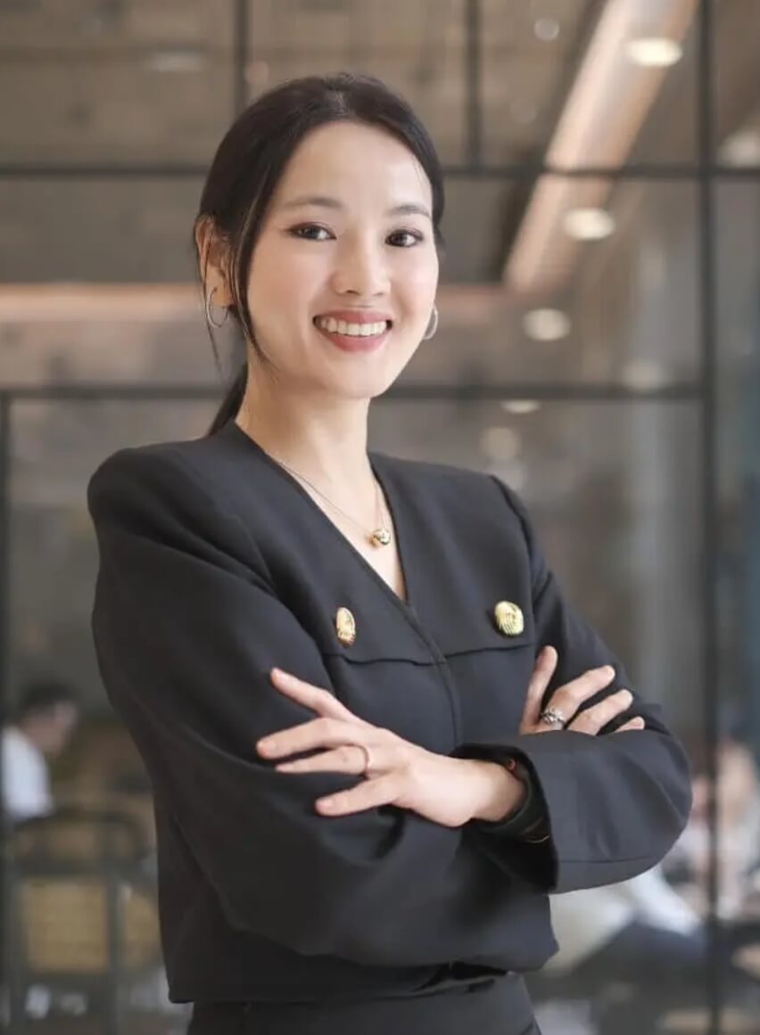 Professional woman in a black blazer with arms crossed, smiling in a modern office lobby.