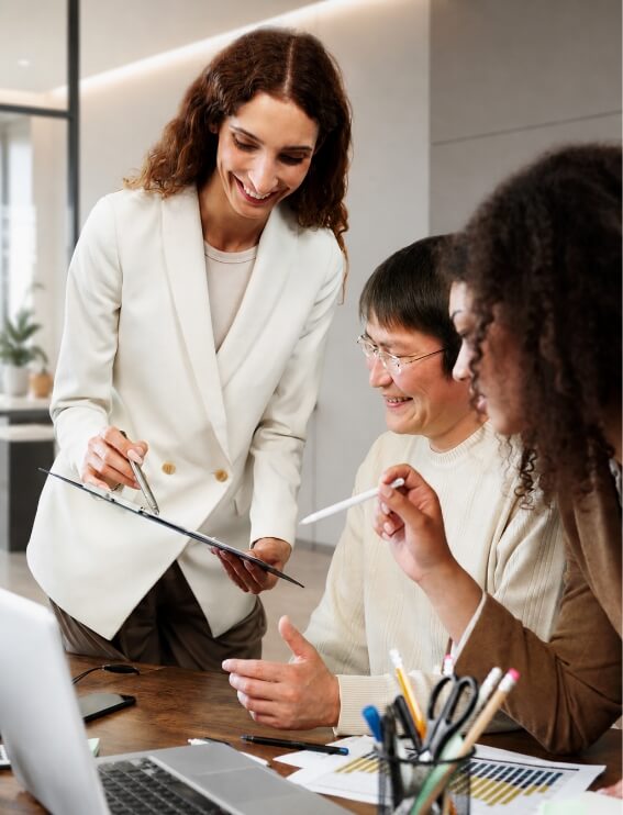 Team of three colleagues collaborating at a desk; one standing with a clipboard presenting to two seated teammates.
