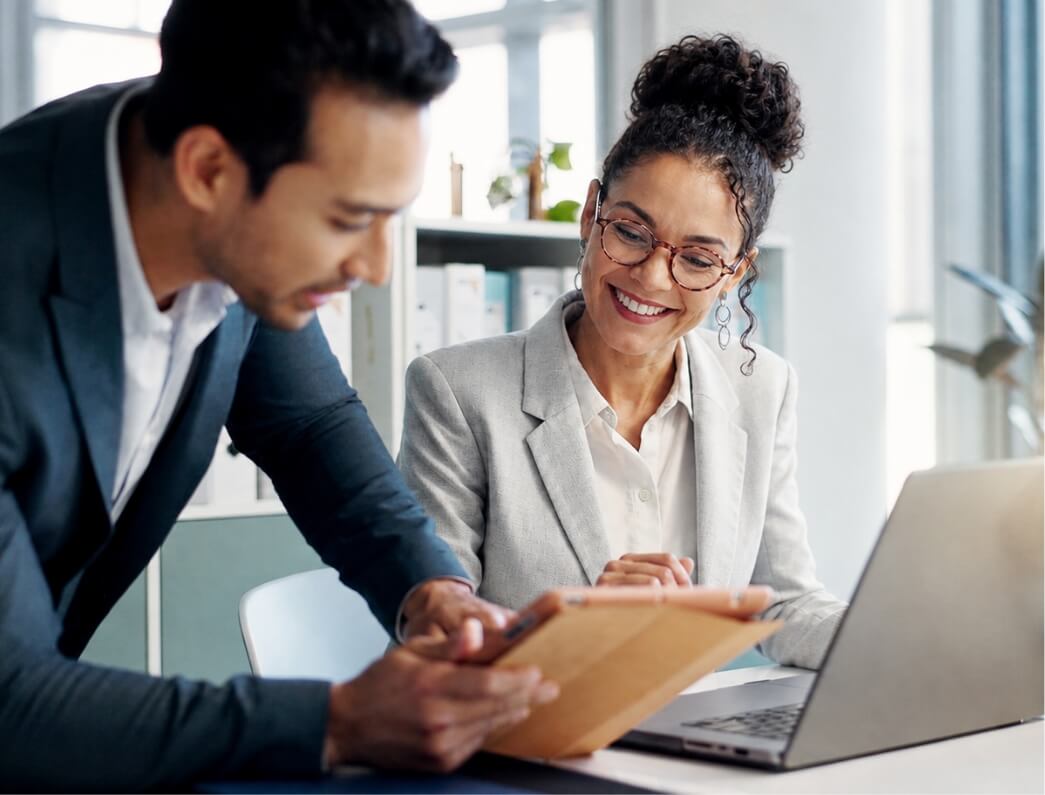 Two colleagues review a document together at a desk, with a laptop nearby.