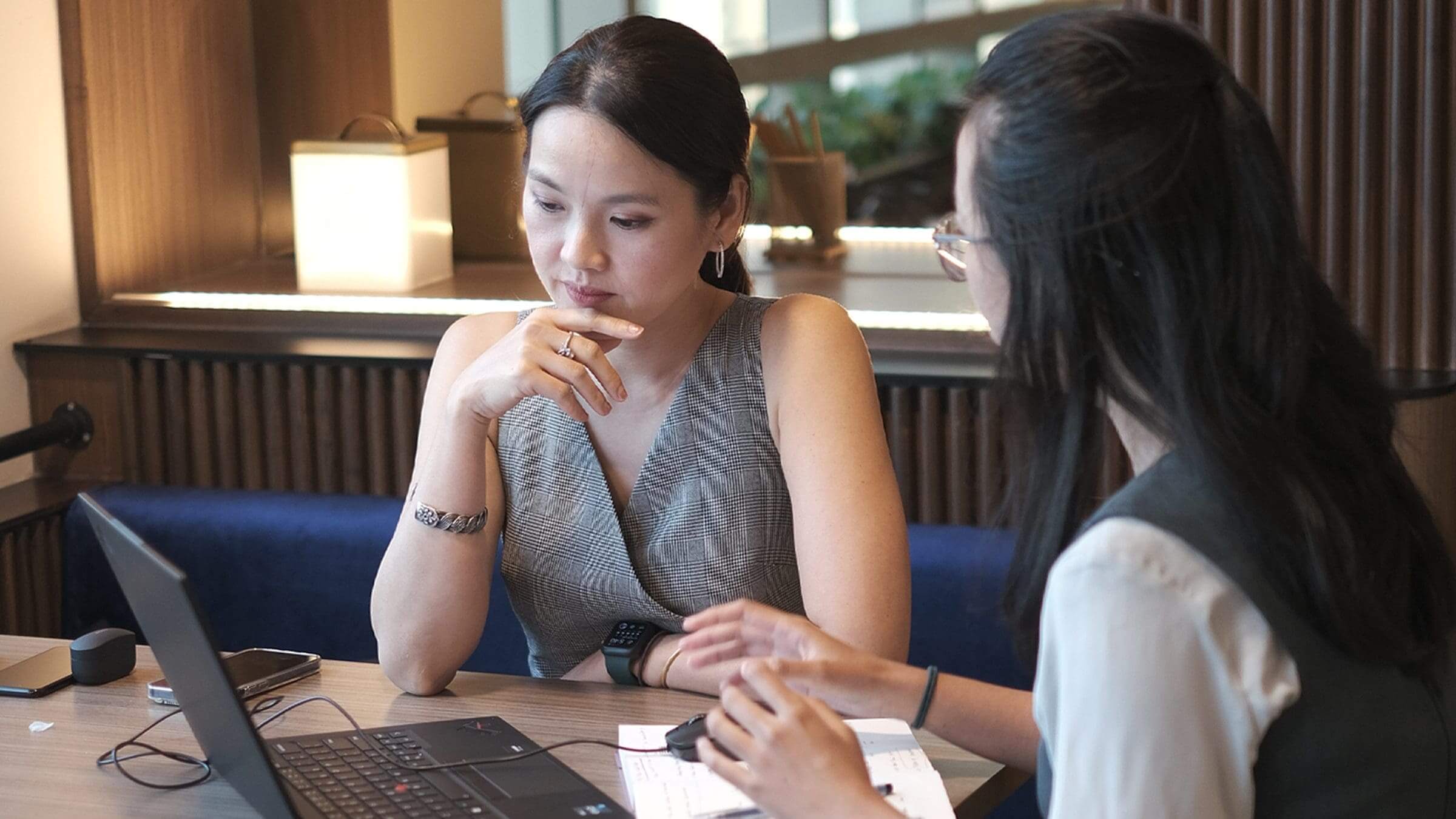 Two colleagues working together at a wooden table with laptops, papers, and a coffee cup in a cafe-style office.