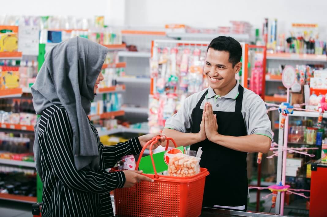 Customer in a hijab hands a red shopping basket to a smiling store cashier at the checkout counter in a bright shop.
