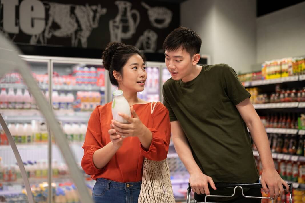 Woman in an orange top shows a bottle to her partner while shopping in a supermarket under bright lights.