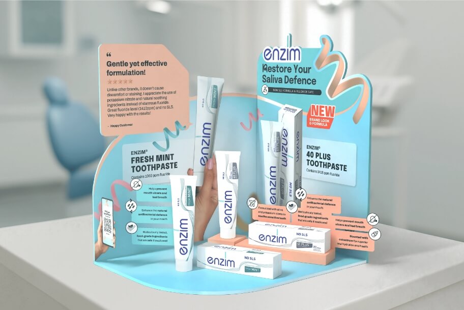 Promotional display of Enzim toothpaste products in blue/orange packaging, featuring Fresh Mint and 40 Plus Toothpaste with slogan 'Restore Your Saliva Defence' in a dental setting.