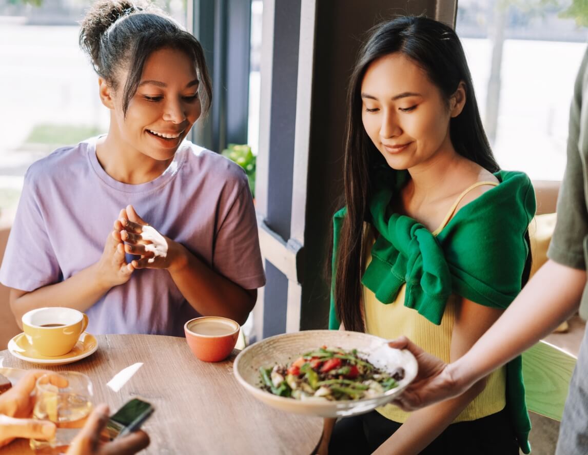 Two women sit at a cafe table; one receives a plate of salad from a server while the other smiles and checks her phone.