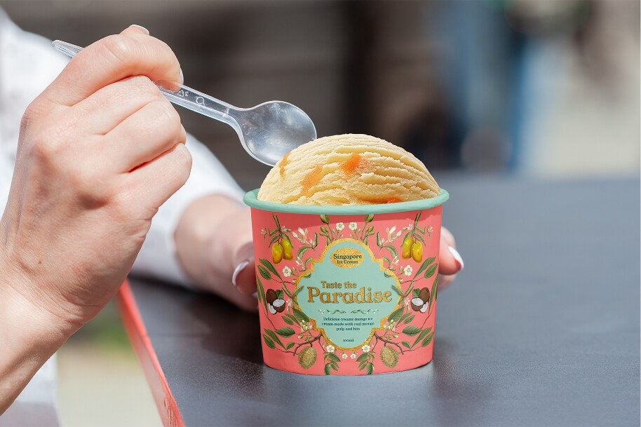 Person holds a pink decorative ice cream cup with an orange scoop and a clear plastic spoon nearby.