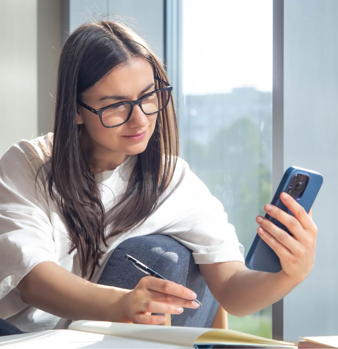 Young woman with glasses sits by a window, looking at her phone while writing in a notebook with a pen.