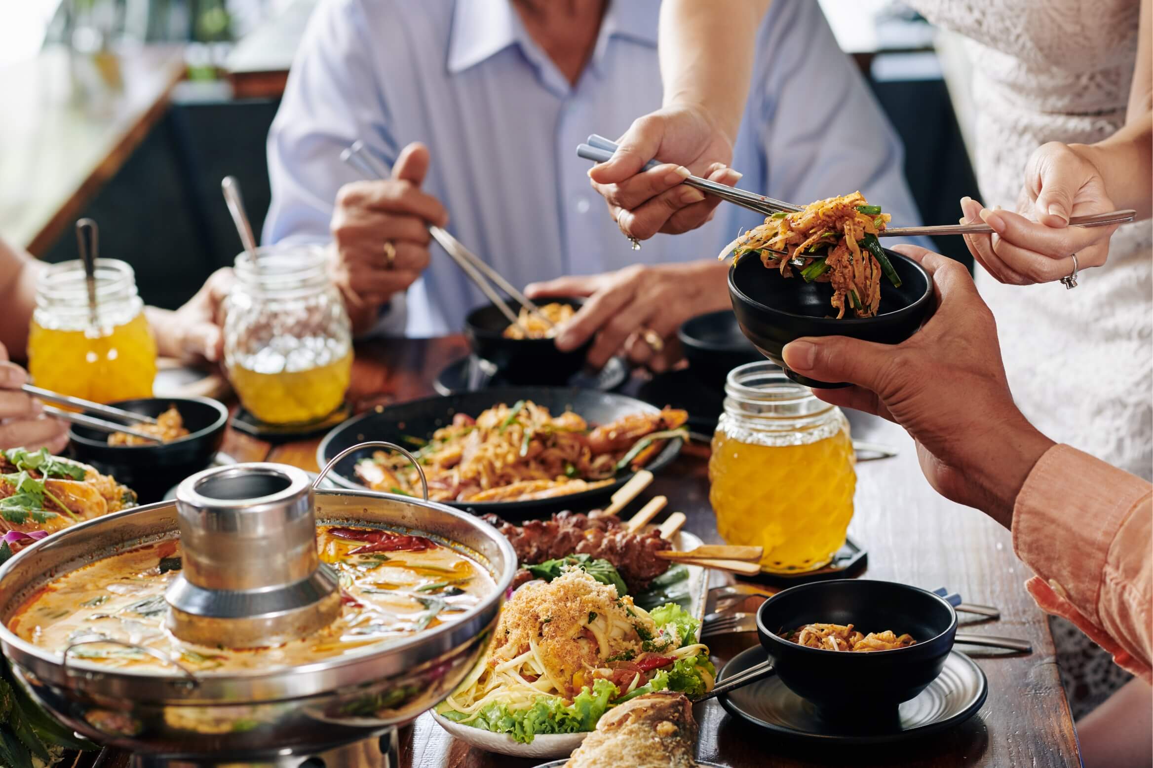 People gather around a table sharing an Asian meal, with chopsticks reaching into bowls and a steaming pot in the foreground.