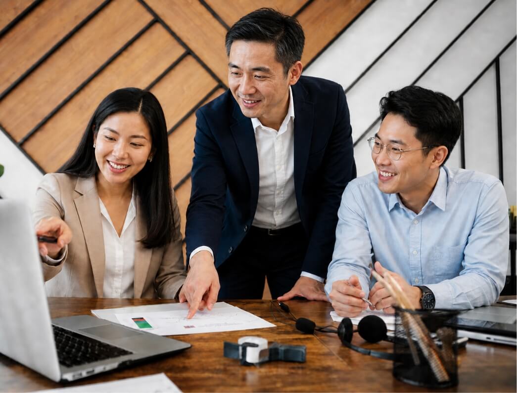 Three colleagues in business attire collaborate around a wooden table, with a laptop and printed documents as they discuss a chart; one person points to the papers while the others smile and listen in a modern office setting.